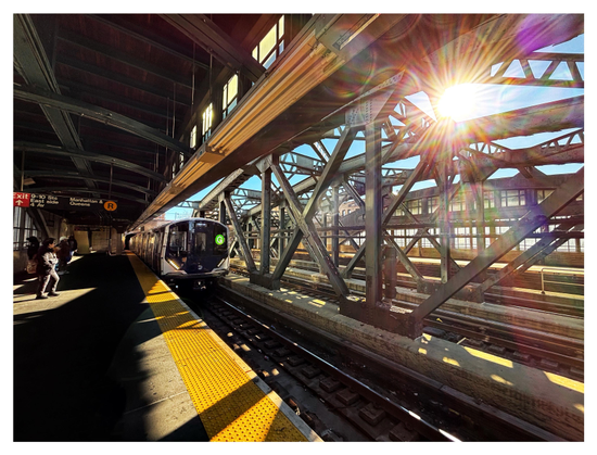Photo taken on the above-ground covered platform of a New York subway station, with the platform and train tracks receding into the distance. Off to the right, the sun shines with a starburst effect through the metal truss over the tracks, illuminating the broad yellow safety stripe on the platform while also casting the truss beams’ shadows onto it. A train with a “G” in a green circle on its front can be seen on the tracks, approaching the camera. The platform is mostly in shade under its canopy, except for a commuter standing in a square of sunlight off to the left. Above the person is a placard with some station information.