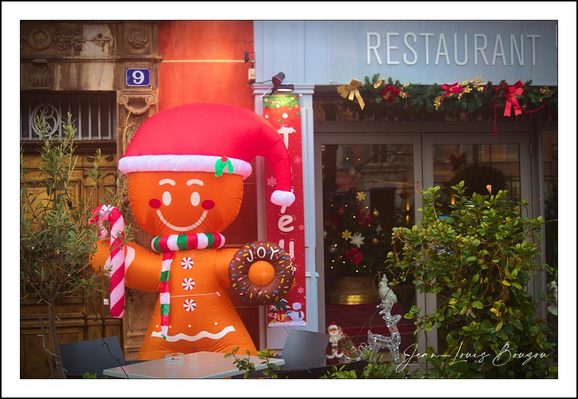 A giant, cheerful inflatable gingerbread person stands sentry outside a small restaurant, spreading an unmistakable holiday vibe. It wears a floppy red Santa hat and a candy-cane striped scarf, its icing smile and rosy cheeks beaming beneath bright, round eyes. One oversized candy cane is tucked into its arm while the other hand holds a sprinkled doughnut-like prop, and white icing zigzags decorate its body like buttons and trim.
Behind it, the restaurant facade is dressed up with evergreen garlands and warm string lights, and potted plants flank the doorway, adding to the cozy, festive scene. The overall color palette — warm reds, oranges and greens — and the soft glow from the decorations make the setting feel inviting and playful, as if the whole storefront were joining in the holiday cheer. A small house number tile and a photographer’s signature in the corner are little, human touches that complete the snapshot.
