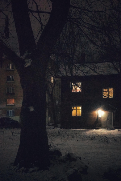 Late evening. The silhouette of a tree is visible in the foreground. In the background is a dark two-storey house with lighted windows and an entrance. There is snow on the ground.  Another house is visible in the distance.