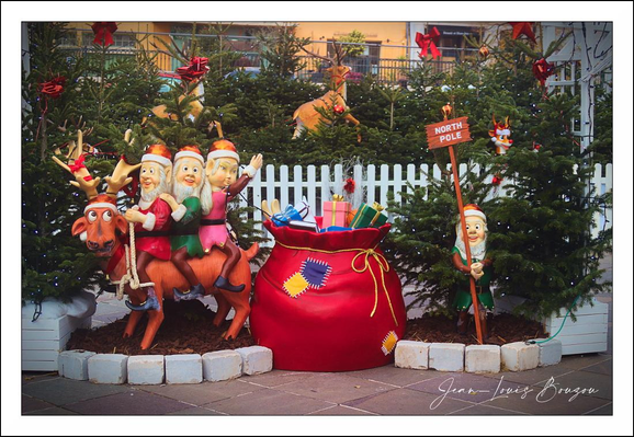 A cheerful, over-the-top holiday display fills the frame: three beaming elf figures in bright red-and-green outfits ride a pair of glossy brown reindeer at the left, frozen mid-prance as if darting off on a toy-delivery mission. In the center sits a huge candy-apple red sack overflowing with colorfully wrapped presents; a blue box and a golden ribbon peek from the top. Flanking the scene are small, heavily decorated evergreen trees tied with big red bows, and a short white picket fence that gives the whole tableau a quaint, village-market feel. To the right a jolly figure holds a wooden sign pointing to the “North Pole,” and little reindeer ornaments and festive lights twinkle in the background. The cobblestone pavement underfoot and the warm glow of nearby storefronts complete the cozy, whimsical Christmas atmosphere.