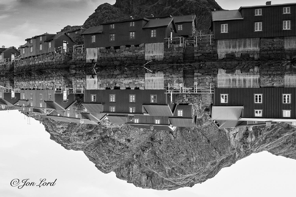 This is an abstract black and white landscape photo in landscape format of a row of Rorbu (Fishermens shacks or cottages) along a harbour waterfront. Stamsund, Norway (2015).

The mirror still waters of the harbour rise upwards from the base to the two-thirds up point in the image. Reflected in the water is a row of restored Norwegian fishermen's Rorbu or Rorbuer, historically used a seasonal housing and storage for fishing equipment, now restored and often used as summer cabins. Here there is a row of four rorbu built along a rock lined harbour wall with a further row of rorbu behind. Inset in the harbour wall are four areas of pale, painted wooden planks laid vertically, perhaps covering previously used spaces for storage or moorings. The Rorbu are rendered dark grey (if in colour, they would be a destinctive dark red or maroon (Falun Red) often associated with Nordic buildings). They are constructed of wood, laid vertically, with six white rectangular windows. The centre section has two floors and a flat roof, at either end there is a single floor extension with a pitched roof. Reflected in the water is a small, rock covered mountain devoid of any trees or vegetation.

The location is the small town or village of Stamsund, in the southern Lofoten Islands, in Norway's arctic north.

I now have to confess: The image is inverted, the harbour water was so still it almost allowed a perfect reflection of the houses and mountain behind. 