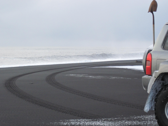 Tire tracks on a black sand beach lead to the back end of a parked vehicle. The tail lights and a shovel attached to the back of the SUV are all that's visible of the vehicle in the foreground. Snow on the beach and white, foamy ocean waves in the background contrast with black sand.