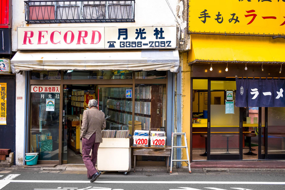 A shabby record shop front. Lines of vinyl shelfs can be seen through the glass. They seem to continue very deep into the shop. A grey-haired fellow is walking in. The sign just says RECORD in all caps, and has a phone number with some Japanese characters above. The Guardian caption: ' In Suginami ward, Tokyo, this old record shop has a striking old sign, and in a city with abundance of such shops, none look quite like this one '