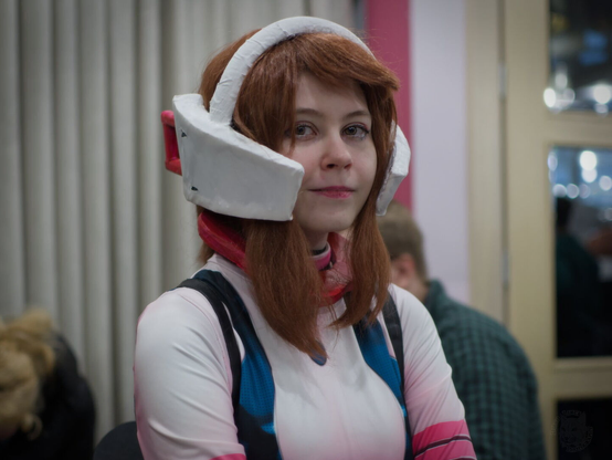 The image captures a moment of quiet contemplation. A young woman with vibrant brown-red hair stands out against the soft, blurred backdrop of what appears to be an indoor setting. She's wearing a white and pink hoodie adorned with blue sleeves, adding a pop of color to her outfit. Overhead, she is also wearing what appears to be a futuristic helmet with big blocks around ears, painted in bold red and white hues. The helmet has an open visor, revealing her focused gaze as if contemplating something significant. Her stance suggests she's standing still, absorbed by whatever lies before her.

The woman's face is turned towards the camera, giving us a clear view of her unique look. In the background, there are two other people partially visible behind her.