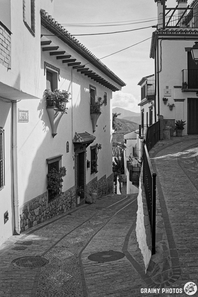 A narrow, winding street in Benelauría featuring whitewashed buildings with decorative balconies and flower pots. The cobblestone path leads up to a scenic view of distant hills under a cloudy sky. Black and white photography captures the quaint atmosphere.