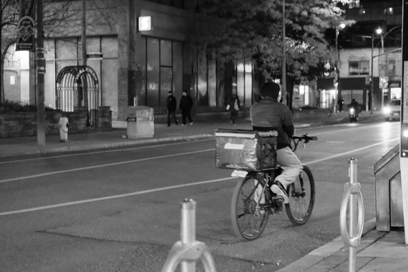 Night outside Wellesley Station. In the background are the buildings across the street, dark but for a few lights. Pedestrians across the street walk towards the left of frame. In the upper right, the lights of cars can be seen coming into frame. Just right of centre, toward the lower edge of the frame, a food courier on a bicycle rights away from the camera toward the right edge of the frame.