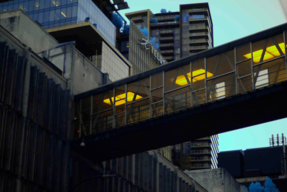 A walkway between two buildings lit in a drab yellow, surrounded by slightly blue-grey buildings, some of which have blue trees on top of them.