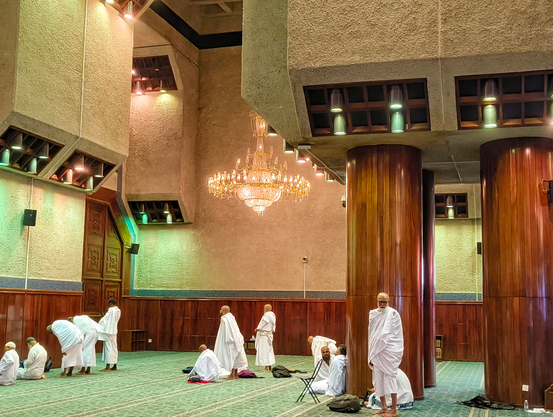 Interior view of a mosque with several people in white ihram clothing. Some are praying on a large green carpet, while others are sitting and resting near large, polished wooden pillars. A large, ornate chandelier hangs from the ceiling, which features textured stone and wooden panels. The lighting is warm and subdued.