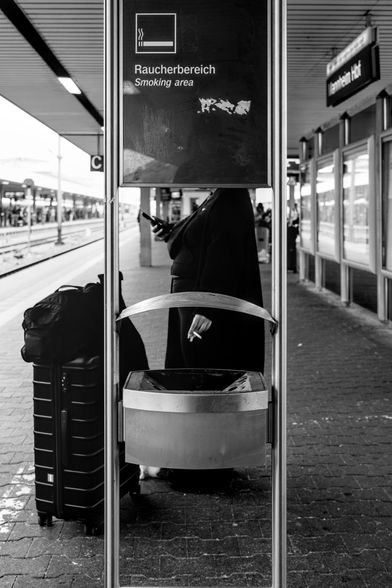 Schwarz-weiß Foto eines Bahnsteigs, eine Person steht rauchend am Handy hinter dem Raucherbereich-Schild, gesucht durch das Schild verdeckt
