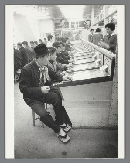 This black and white photograph captures a group of individuals engaged in playing slot machines. The focus is on the participants, who appear to be men based on their attire, such as hats and jackets that suggest an earlier era's fashion possibly from mid-20th century Japan due to cultural elements like kimono-like clothing for some players.

The room has multiple rows of gaming booths with visible slot machine designs. The patrons are seated at the machines, interacting with them by inserting coins or tokens into slots and pulling levers. A woman behind a counter is overseeing their activities, possibly as an attendant ensuring smooth operation or enforcing rules.

The environment suggests a public place designated for gambling, likely within a larger venue like an arcade hall or casino space in Osaka's Kamagasaki district during the year 1960. The lighting is bright and artificial typical of indoor settings from that period. There are no modern electronic devices visible; only mechanical slot machines which were prevalent at the time.

This scene reflects leisure activities and social interactions centered around a form of gambling, providing insight into entertainment practices in Japan's mid-20th century urban culture as documented by photographer Ed van der Elsken.