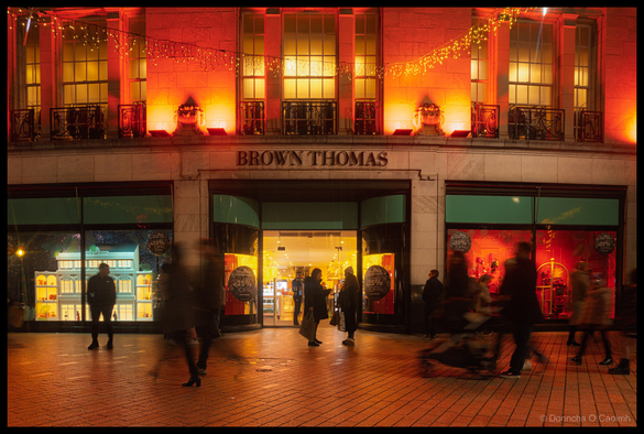 Night-time photograph of Brown Thomas storefront on Patrick's Street, Cork, with dramatic red and orange architectural lighting illuminating the upper facade featuring ornate iron railings and window frames with festive wreaths, illuminated shop windows below displaying promotional signage reading "25%", pedestrians in silhouette walking on brick pavement.