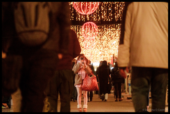 Night-time photograph of pedestrians walking on Oliver Plunkett Street, Cork, beneath cascading golden fairy lights creating a canopy effect overhead, large red neon-illuminated festive spheres and decorative motifs suspended above the street, blurred figures in winter clothing in motion, storefront signage visible at street level.