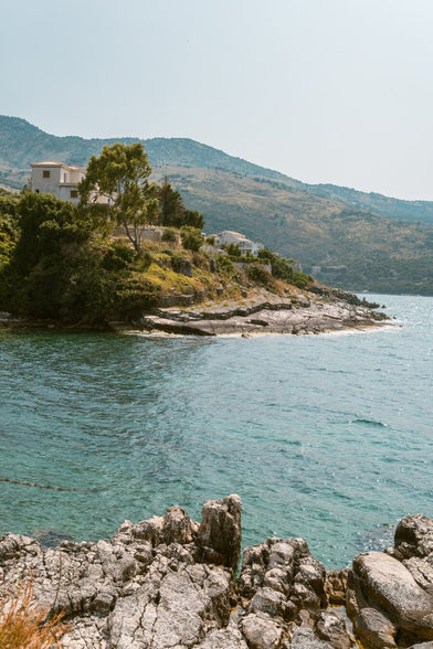 A photo of a bay with rocky beaches, buildings and trees visible in the background. The sky is cloudless.
