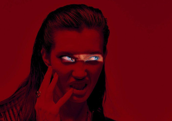 A figure in a black leather jacket illuminated by a light beam across her blue eyes stands against a deep red background, exuding a bold, mysterious vibe.