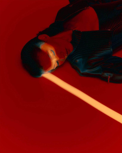 A model in a sleek black jacket lies on a vibrant red background, illuminated by a striking yellow light beam across their forehead.