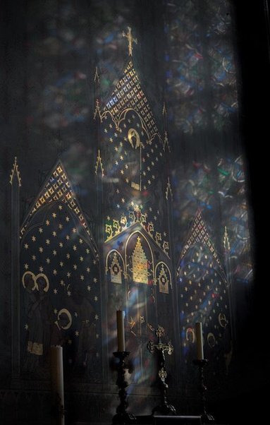 Photo d’un mur à l’intérieur de Notre-Dame de Paris, décoré de dorures représentant une cathédrale stylisée, frappé d’un faisceau lumineux coloré venant du vitrail sur la droite