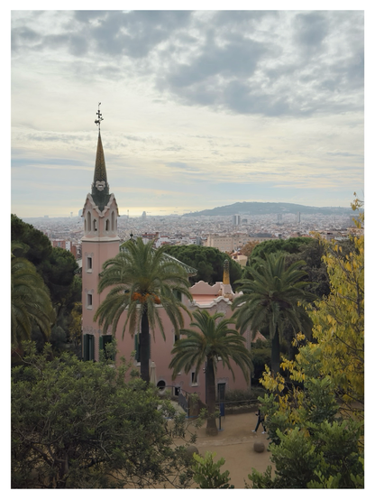 A whimsical, pastel-pink building with a tall, tiled spire—characteristic of Antoni Gaudí’s architecture—stands among lush palm trees in Park Güell. In the background, Barcelona stretches toward the hazy horizon, with the sea faintly visible under a bright, cloud-streaked sky. Trees frame the foreground in varying greens and yellows, adding depth to the scene.