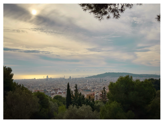 A panoramic view of Barcelona framed by trees, with the sun glowing through hazy clouds above the city and the Mediterranean shimmering on the horizon.