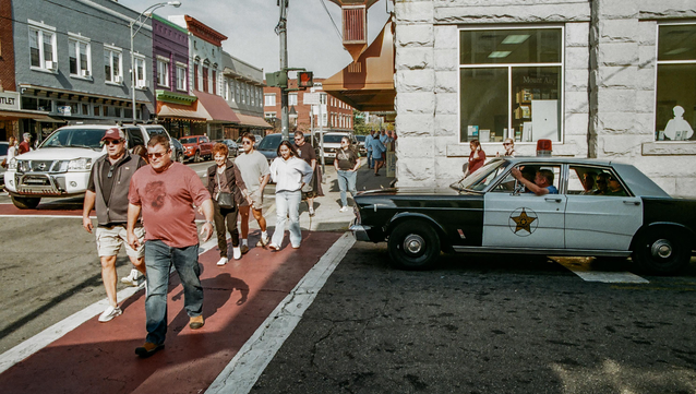 A group of folks crossing the street while an antique police car full of people is waiting to turn on to the main street.