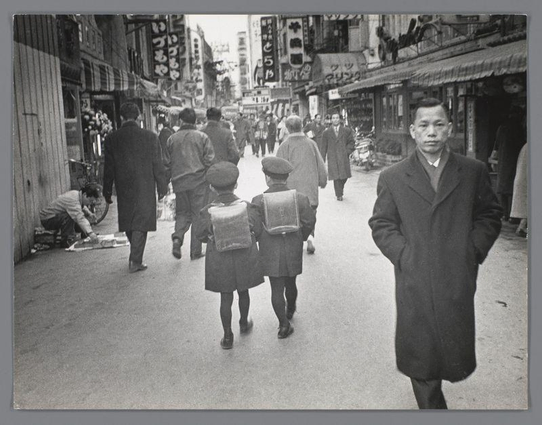 This black and white photograph captures a bustling street scene in Osaka, Japan during the 1960s. The image shows various individuals engaged in daily activities: some are walking along the sidewalk, while others appear to be shopping or waiting at stalls that line both sides of the busy thoroughfare. Two children with backpacks on their backs walk ahead of a man dressed in a coat and trousers, who is looking towards the camera. Their attire suggests it might be cool weather.

The architecture reflects an urban environment typical for Japan during this period: tall buildings adorned with signs featuring Japanese characters dominate the scene. The signage indicates commercial establishments such as shops or restaurants, adding to the lively atmosphere of the street depicted in Ed van der Elsken's photograph from 1960.