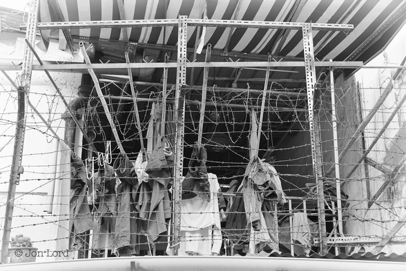 This is a black and white photo of a load of washing drying on a balcony surrounded by barbed wire. Kampong Glam, Singapore (2015).
The camera is looking upwards slightly from the street at a first floor balcony of a rough looking house / very low-rise and low rent, apartment building. There is no window or door behind the balcony, just a wide opening with a dark interior inside. Surrounding the balcony is a Dexion steel strip framework about a metre of a half tall, supporting thirteen horizontal runs of barbed wire with a coil of barbed wire on top. Over the balcony is an black and white striped awning sloping downwards gently from the building towards the edge. Stretching from the inside to the edge of the balcony are seven bamboo poles each with laundry resting on then and drying in the sun.