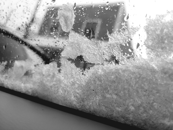 Black and white photograph of a car window covered with snow and water droplets, with a blurred view of the outside visible. The lower section of the window is coated with thick, textured snow, while the upper section has water droplets and some snow patches. A dark, diagonal line appears in the lower-left corner under the car's window. Through the window, a blurred cityscape can be discerned.