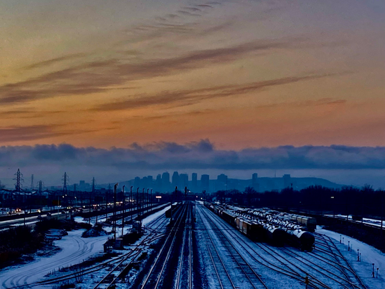 Railroad tracks in the foreground lead from a train yard to the Montreal skyline on the horizon. The island appears to be against a backdrop of white mist, under a low cover of clouds. Above the clouds, the sky is orange and yellow with purple streaks of clouds. 
