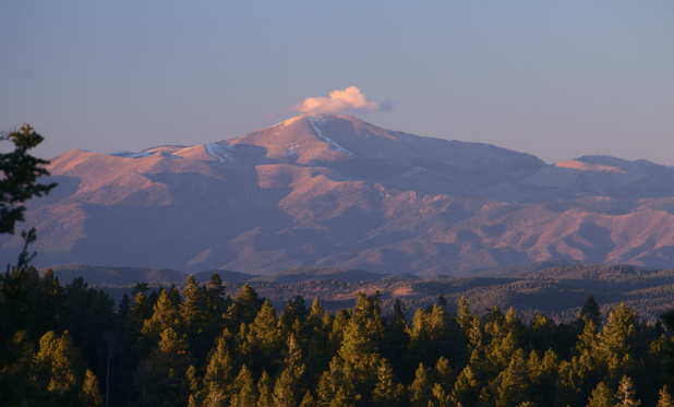 Sierra Blanca's upper flanks and slopes are devoid of vegetation, and show a few traces of snow. The low sun angle accents the terrain's relief. Conifers cover the foreground ridge a quarter mile away, and also cover distant terrain at the base of the mountain. A small cap cloud hangs just above the summit, otherwise the sky is blue. The overall light is reddish-warm because we are close to sunset. Nearby shadows are deep and contrasty. Distant shadowed terrain is lower contrast due to haze.
