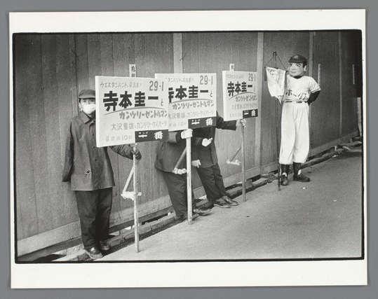 The image is a black and white photograph depicting four individuals standing against a wall. Two of them are holding up signs with Japanese text, which appears to be advertising or promotional material related to the year 1961 as indicated by "29-1" on each sign. The person in the center is wearing a baseball uniform with the number '73' and has his mouth open wide as if shouting or calling out something energetically.

To their left, one individual wears a mask over his face, suggesting health precautions might be necessary at this time period, possibly during an outbreak such as the flu. The other two individuals are dressed in casual clothing suitable for urban work environments. They stand with hands on hips and seem to be promoting or supporting whatever is advertised by the signs.

The setting appears to be a street next to a building, given the wall behind them and what looks like part of a bench visible at the bottom left corner. The ground shows some litter around their feet. There's no direct sunlight casting shadows here; it suggests an overcast day or shaded area typical for urban environments in Japan during that era.

This photograph is captioned "Mannen met reclameborden en een masker, Osaka" which translates to "Men with advertising boards and a mask, Osaka." The photographer of this image is identified as Ed van der Elsken from the provided source link.