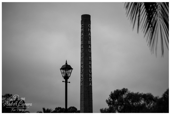 Full colour photograph of the main facade of Chateau Tanunda, featuring symmetrical red brick and stone work, arched windows, and dark sloping roofs.

A wide, bright green lawn and a central gravel pathway lead the eye toward the grand entrance under a bright blue sky with wispy white clouds.