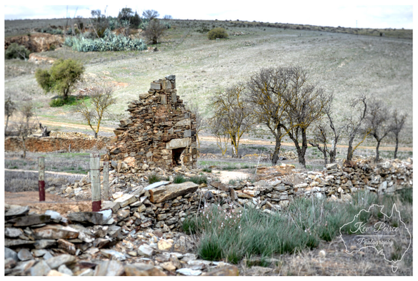A photo of the stone remnants of a ruin in a dry, open field. A section of a stone wall and the corner of a collapsed structure are prominent in the foreground.

Several bare-branched trees stand behind the ruins, and a dusty hill rises in the background.