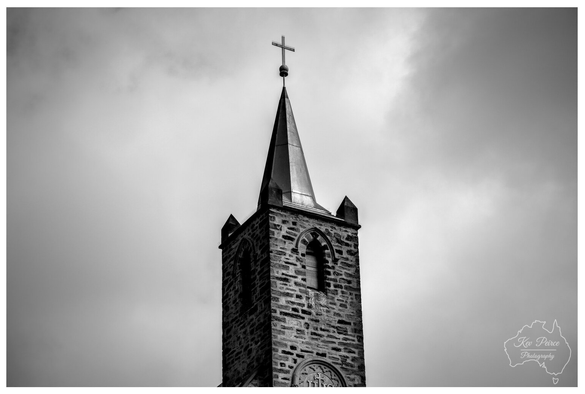 Black and white photograph showing the tall, stone steeple of a church in Burra, South Australia, topped with a cross.   The spire is dramatic against a cloudy, atmospheric sky, emphasizing the architectural details of the bell tower.