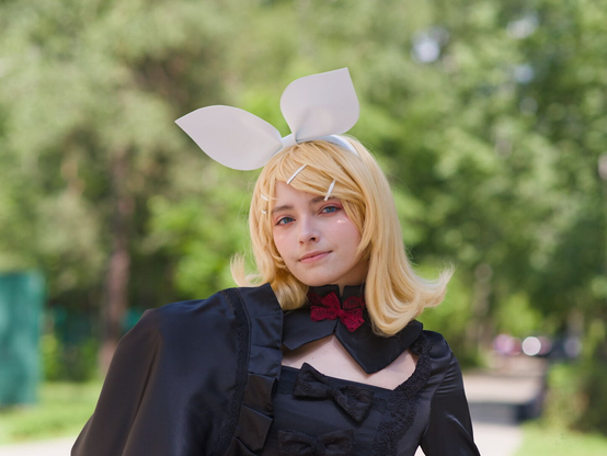 The image captures a person dressed in an elaborate costume, standing outdoors. They is wearing large white bunny ears on their head and has blonde hair styled with bangs that fall over the eyes. The attire includes a black outfit adorned with intricate lace detailing around the neck and shoulders; it features a prominent red bow at the collar which adds contrast to the ensemble. Their expression is serious, yet slightly playful, aligning well with the whimsical nature of their costume.

In terms of location, there appears to be greenery in the background suggesting she's near trees or bushes. The daylight suggests this photo was taken during a time when natural light can illuminate outdoor scenes effectively without harsh shadows.

This image could potentially have been captured at an event where costumes are worn such as a cosplay convention. It showcases the costume in detail and captures the playful yet serious mood of someone ready for such events.