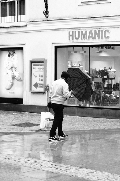 Das Foto zeigt eine Straßenszene in Schwarz-Weiß in der St. Pöltenter Innenstadt an einen regnerischen windigen Tag. Man sieht eine Person mit Regenschirm der durch den Wind leicht verbogen ist und die Person den Regenschirm nicht übern Kopf hält sondern davor. 
Im Hintergrund die Auslage von Humanic zu sehen.