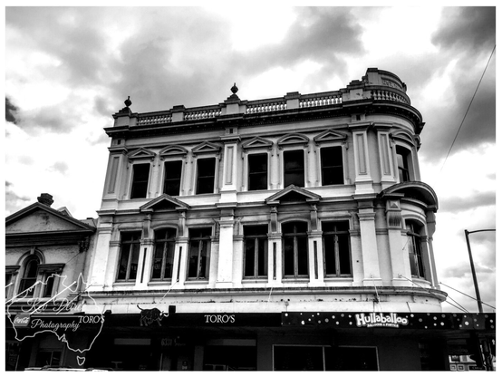 Black and white photograph of a stately, three story Victorian era building in Launceston, Tasmania, featuring arched windows and a prominent corner turret with a balustrade roofline against a cloudy sky.  The ground floor businesses include TORO'S and Hullaballoo.