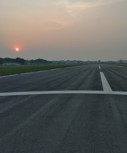A sunset over an empty runway, casting a warm orange glow across the sky. The straight path of the asphalt leads towards distant silhouettes of trees and buildings.