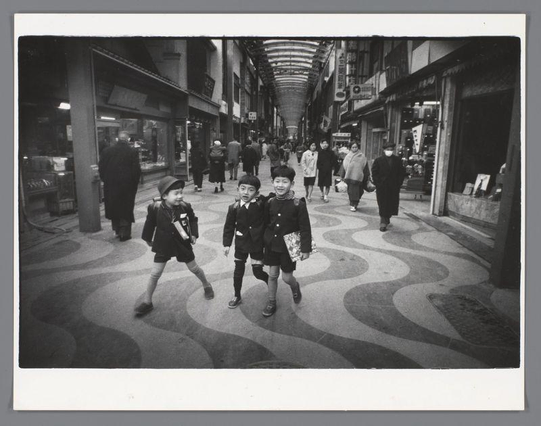 The image depicts a bustling urban street scene in black and white, likely from the mid-20th century judging by clothing styles. Two young children are at the center of the frame, walking away from the camera with one arm around each other's shoulders; they appear to be siblings or friends. The boy on the left wears dark school uniform trousers paired with a lighter jacket and is carrying books in his hand, while the girl beside him has short hair and also sports a similar style of dress typical for children at that time.

Behind them are several adults walking in various directions, including what appears to be an elderly person wearing spectacles. The background shows a series of shops along both sides of the street with storefronts displaying goods such as hats or scarves on mannequins. Above the pedestrians is an arched ceiling supported by columns that create a patterned effect on the ground below.

The overall atmosphere suggests everyday life in a Japanese city, possibly Osaka given the context provided and the style of clothing which seems to reflect late 1950s or early '60s fashion trends found within historical records. The image captures candid moments with no apparent staging, indicating it was likely taken spontaneously as part of daily activity rather than for artistic photography purposes.

The photograph is credited to Ed van der Elsken and the provided URL suggests that this particular work can be resear [...]