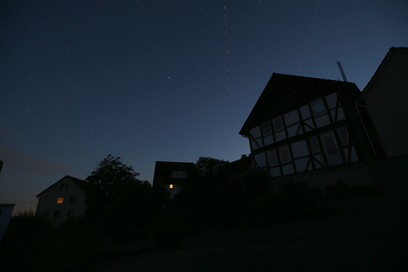 Foto, Dorfszene bei Nacht: rechts ein Fachwerkhaus; am fast klaren Himmel viele Sterne mit leichter Erdrotation, dazu ca. mittig und fast senkrecht die Spur der ISS.