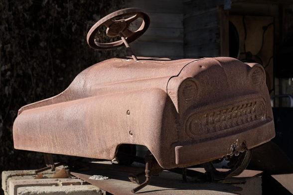 A color landscape photo of a small kid's metal petal car toy. The car faces toward the right. The right side is in bright sunshine and the front in shadow. There are no wheels and it sits on its axle. The front of the rusty car it looks like a grimacing face with wide eyes and clinched teeth.