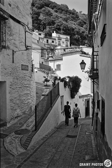A narrow, winding street in Benelauría, flanked by whitewashed buildings with traditional architecture. Two people stroll down the cobblestone path, surrounded by greenery on the hillside above. A vintage street lamp adds charm to the scene.