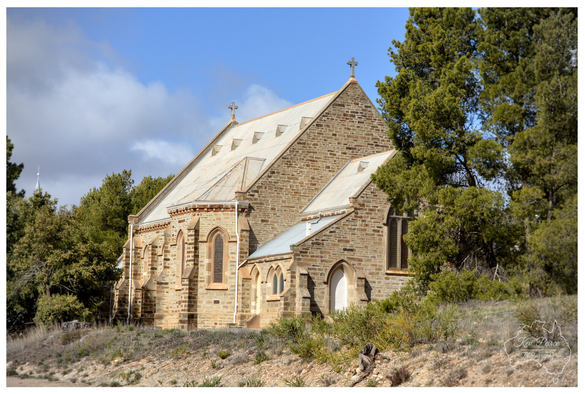 Photograph showing a historic stone church building on a slight incline under a bright blue sky with white clouds.  The church features Gothic style windows, a corrugated iron roof, and is flanked by tall, dense green pine like trees and dry scrub in the foreground.