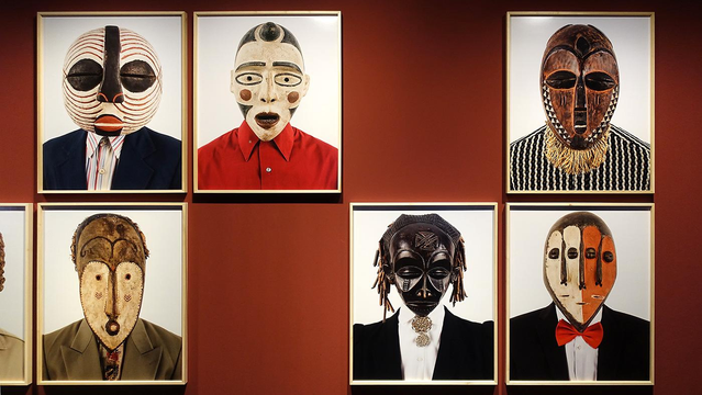 Rode muur van het museum met daarop in twee rijen boven elkaar allemaal portretten van mensen met verschillende Afrikaanse maskers in kleur, vorm en uitvoering.