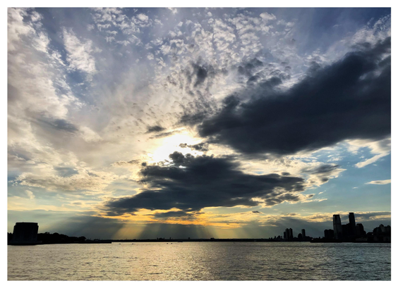 Photo of a wide bay with calm waters at sunset. Building silhouettes sit at the horizon to the left and right of the image. Clouds of different shapes fill the sky, in particular two shadowy masses partially obscuring the sun, which sends out dramatic golden crepuscular rays over the water.