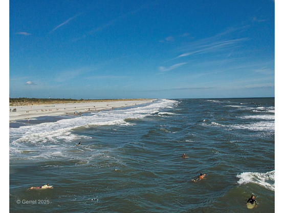 Aerial view of a sandy beach with gentle waves under a clear blue sky. Swimmers and surfers are scattered in the ocean, conveying a peaceful, sunny day vibe.