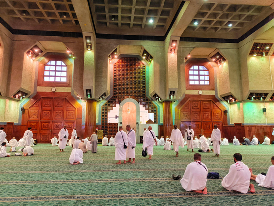A wide-angle photograph inside a large mosque, showing numerous pilgrims dressed in white Ihram garments standing and sitting on a sprawling green carpet. The mosque features a high, coffered ceiling with square recesses, large wooden doors, and geometric patterned window screens. The architecture is modern with light-colored stone or concrete walls and warm wooden accents.