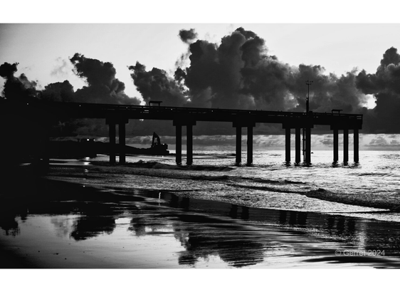 Silhouetted pier against dramatic clouds at sunrise, reflected on the wet sand. The calm ocean and sky convey a serene, contemplative mood.