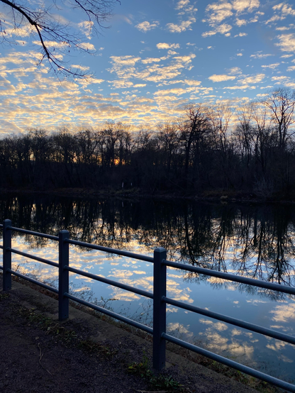 A scene close to a river.
In the foreground there‘s a metal railing on a foodpath alongside a river.
Beyond the railing the blue sky full of white clouds and the trees are being mirrored in the water of the river.
On the other shore (background) there are the black silhouettes of trees growing there.