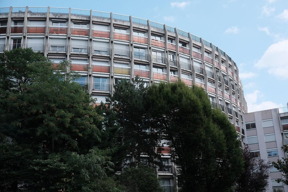 Une partie d’un immeuble circulaire, avec des barrière de balcon alternant entre une grille et une plaque de verre rouge sombre entre chaque étage