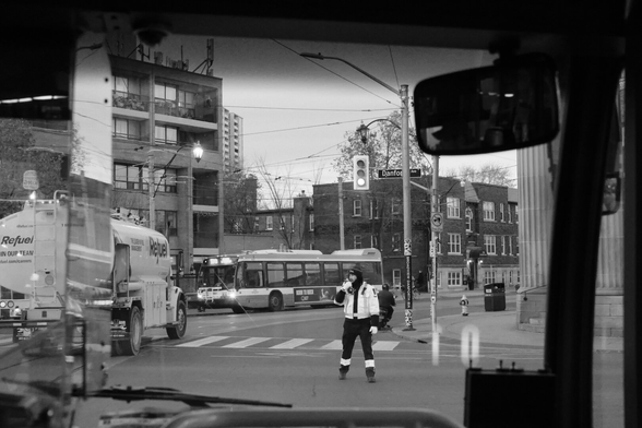 Shot through the front windshield of a bus, we see a busy intersection. There’s a tanker truck at left and another bus in centre background. Just right of centre in the middle of the intersection is a cop directing traffic, his safety gear standing out against his surroundings.