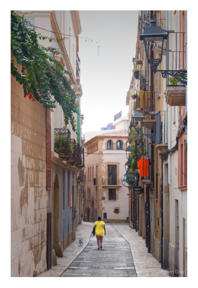 View of a mediaeval street with lanterns and balconies. From a balcony hangs a red dress. A woman in a yellow dress is seen from behind. She is walking a small dog on a lead.